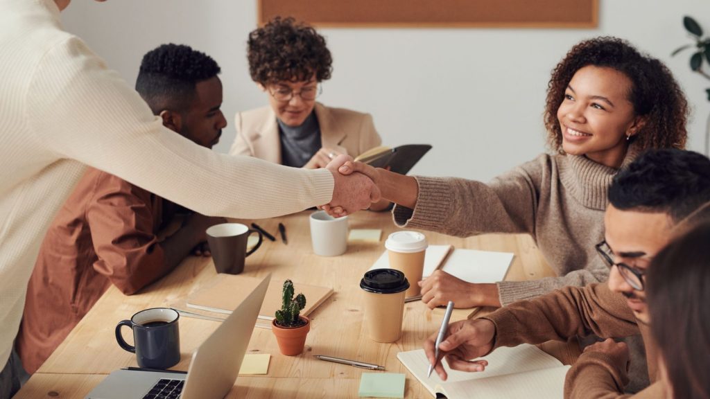 A diverse team welcoming a new colleague with a handshake around a meeting table